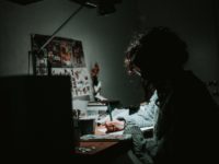 low light photography of woman in gray knit sweatshirt writing on desk