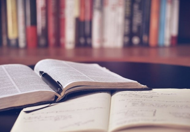 Imagem de livro e caderno abertos sobre mesa, com livros em segundo plano em estante